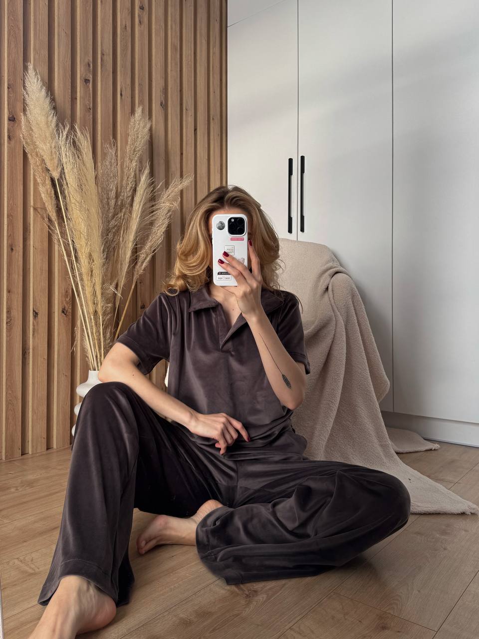 Person taking a mirror selfie in a dark outfit with a phone, sitting on the floor in a room with wooden walls and a white cabinet.