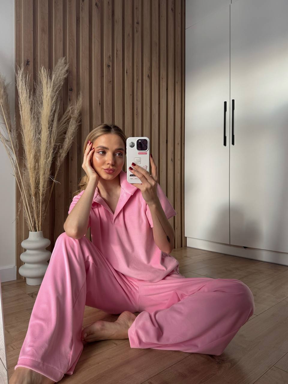 Woman in pink pajamas taking a selfie in a room with wooden floor and white wardrobe.