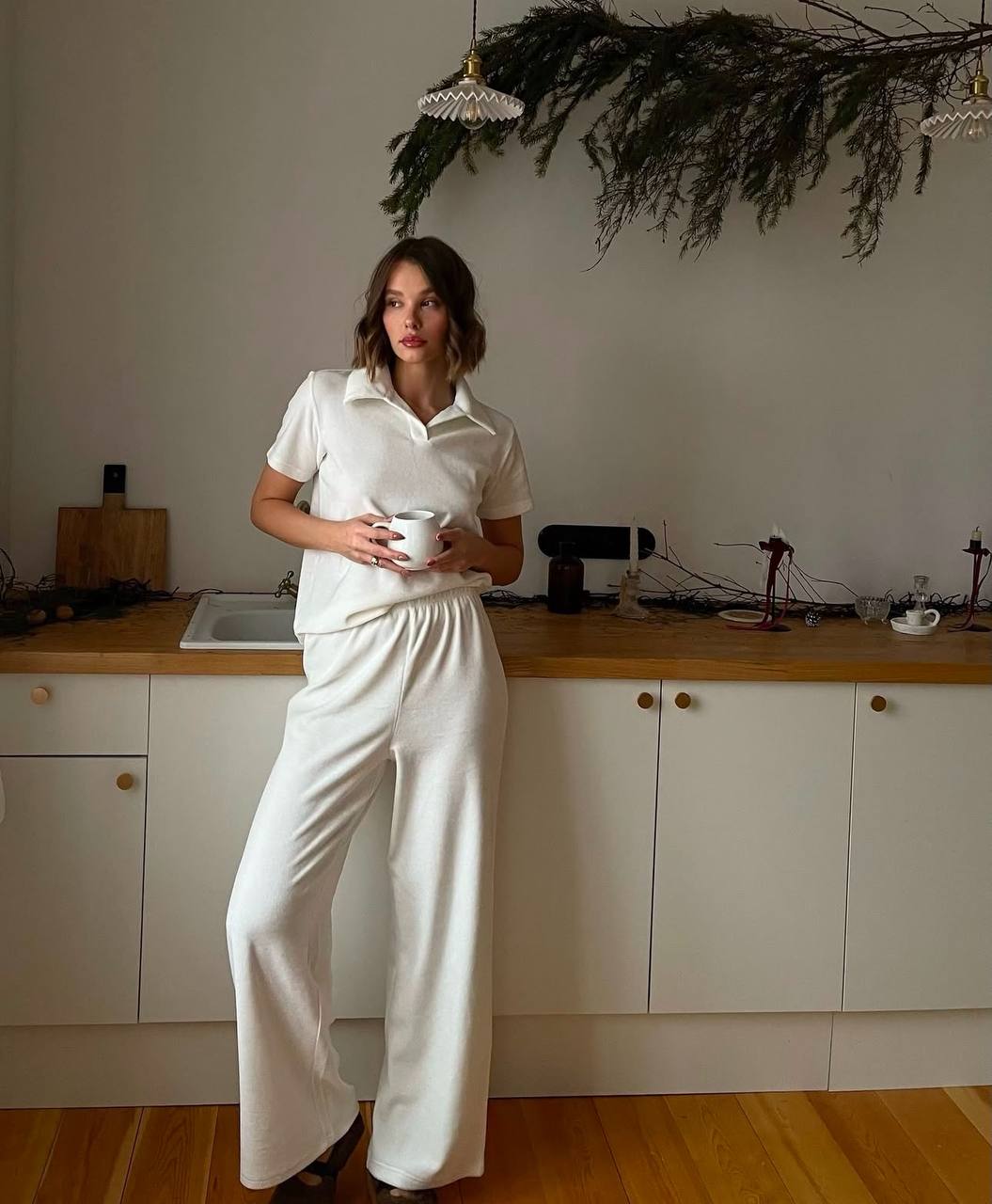 Woman in a white outfit standing in a kitchen holding a cup.
