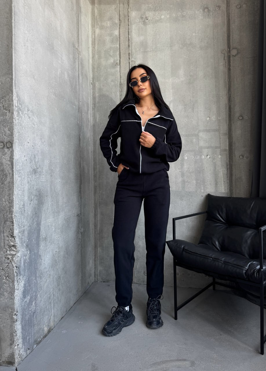 Person wearing a black tracksuit standing against a concrete wall with a black chair.