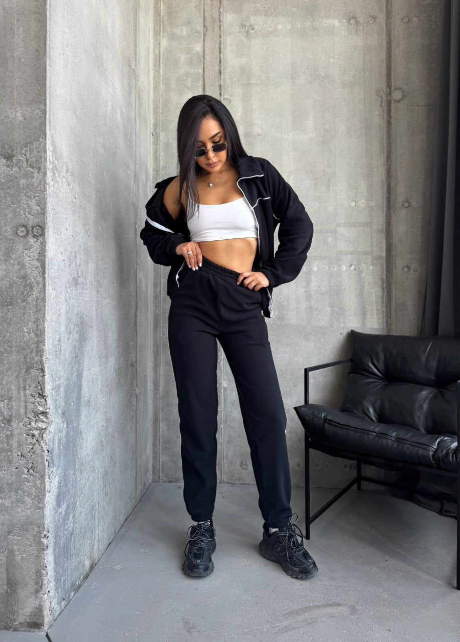 Woman in black jacket and pants standing against a concrete wall with a black leather chair.