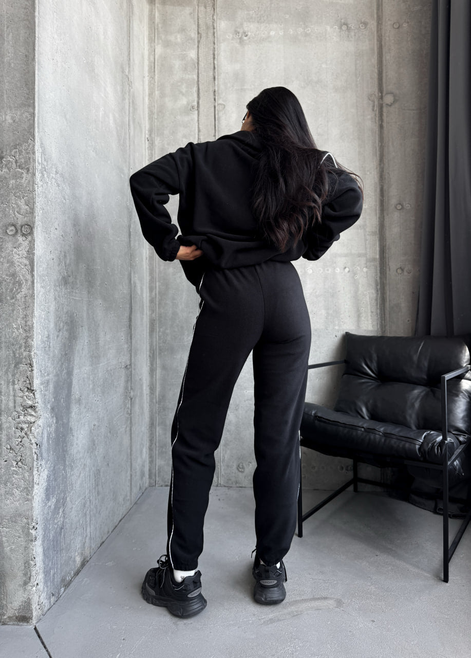 Person wearing a black tracksuit standing against a concrete wall with a black leather couch.