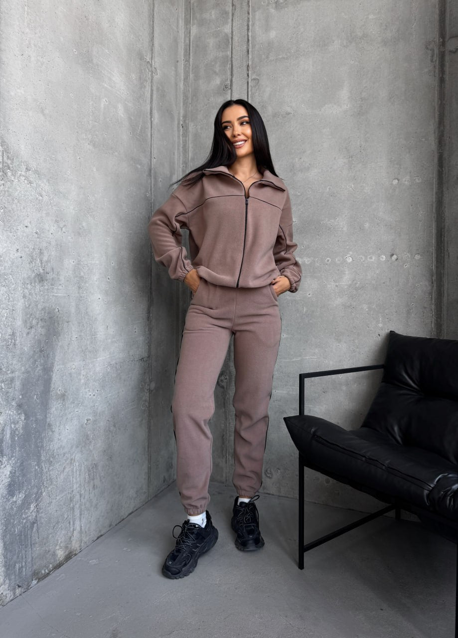 Woman in a brown tracksuit standing against a gray concrete wall with a black chair.