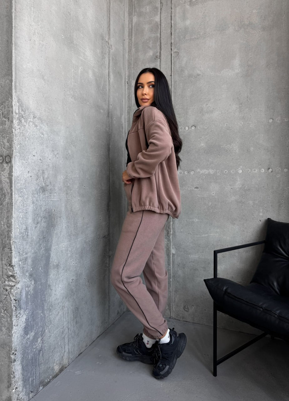 Woman in a pink tracksuit leaning against a gray wall with a black chair in the corner.