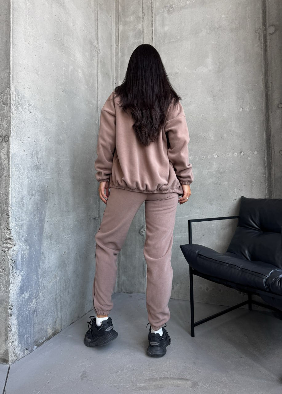 Person wearing a beige tracksuit standing against a concrete wall with a black chair.