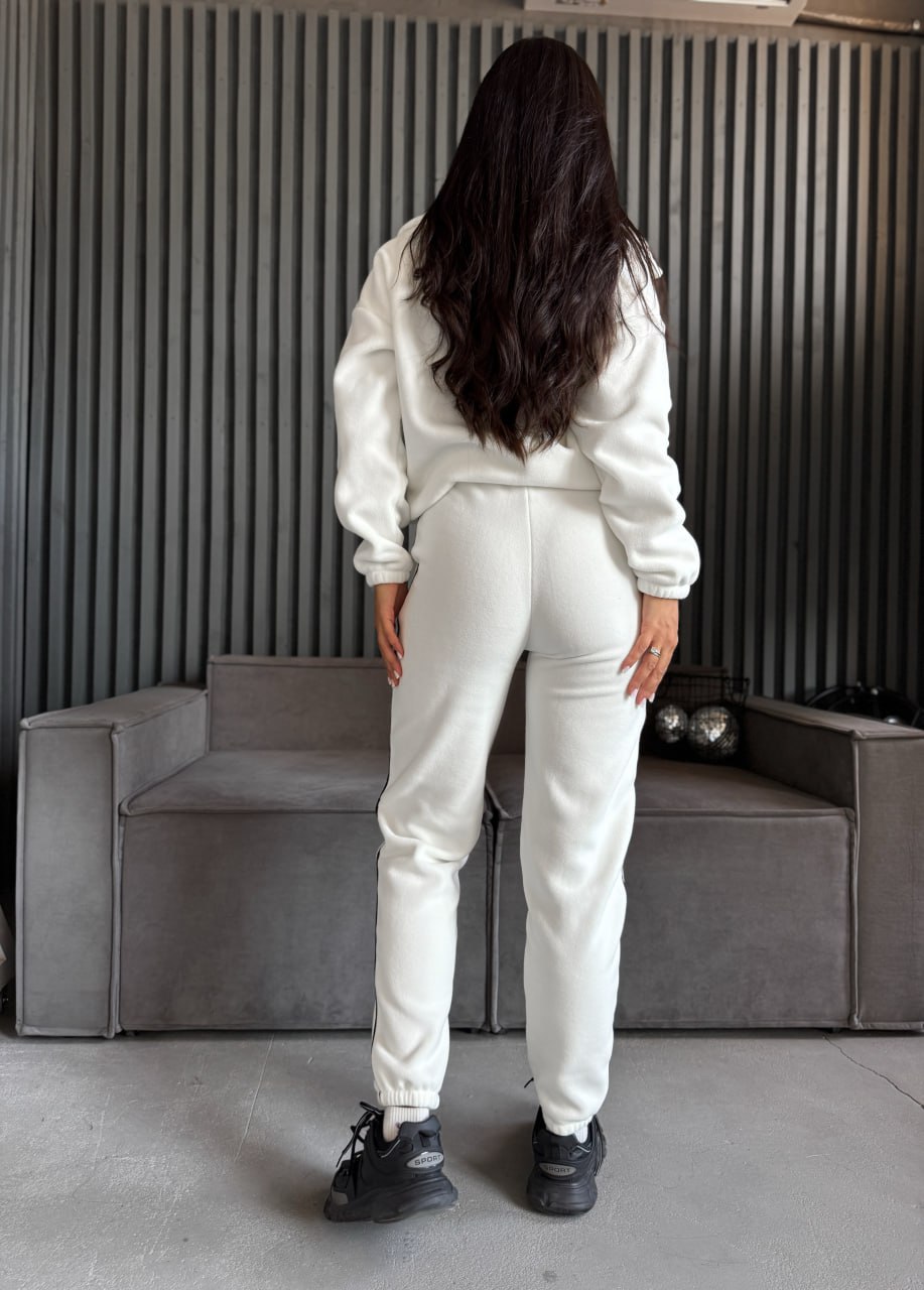 Person wearing a white tracksuit standing in a room with a gray couch and striped wall.