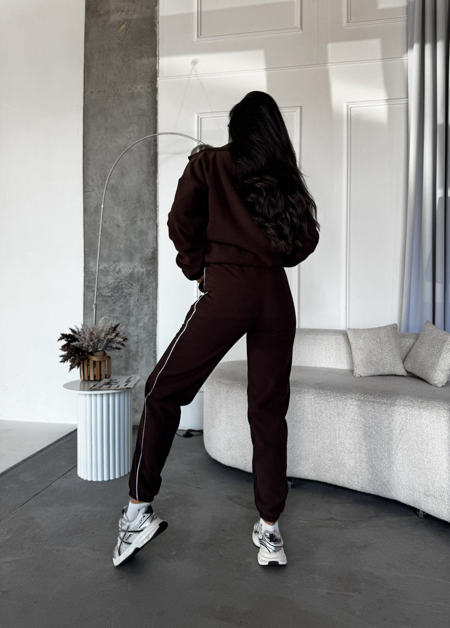 Person wearing a dark brown tracksuit in a modern room with a sofa and decorative items.