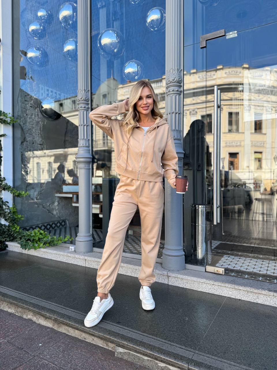 Woman in a beige tracksuit standing outside a building with reflective glass doors.