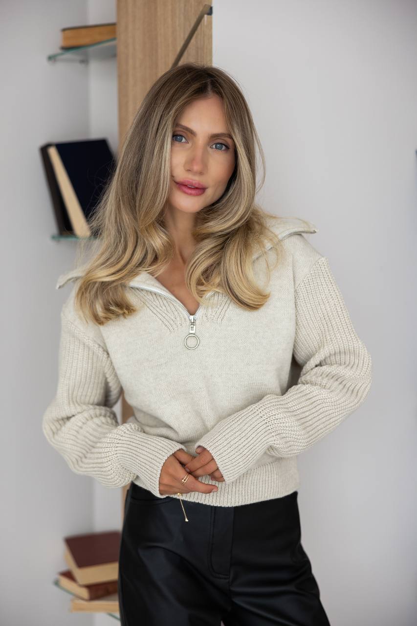 Woman wearing a beige sweater with a white shirt underneath, standing against a light background.