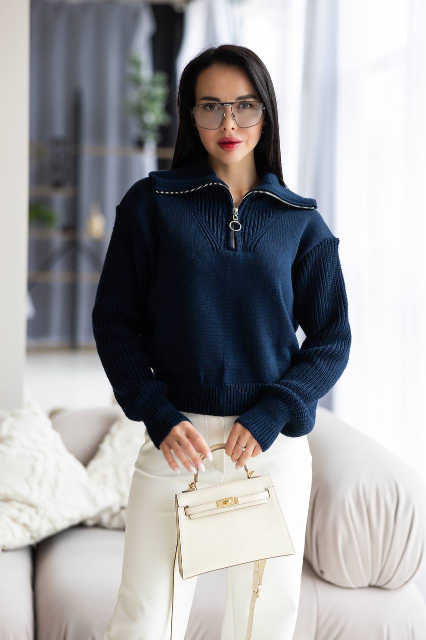 Woman wearing a navy blue sweater and white pants holding a cream handbag indoors.