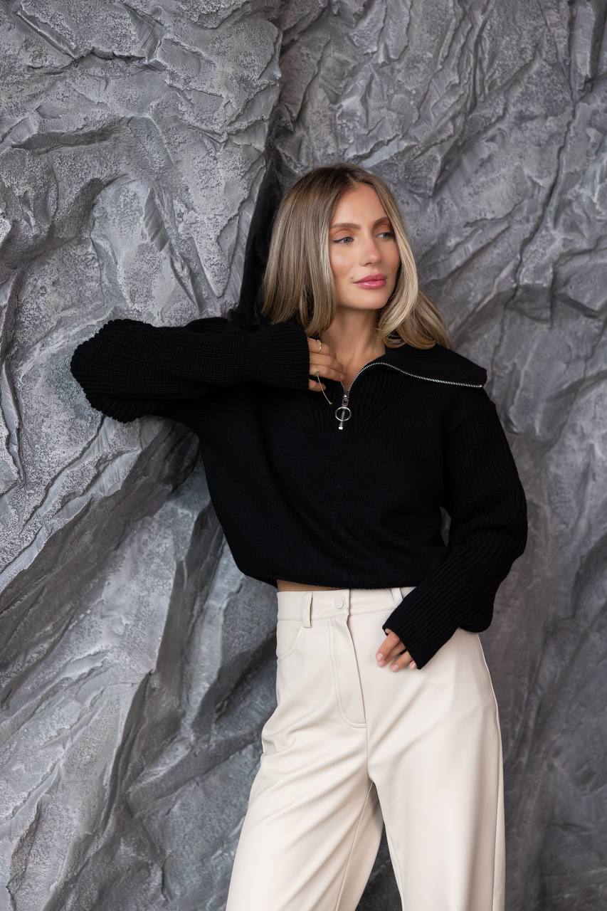 Woman wearing a black sweater and white pants against a textured gray wall