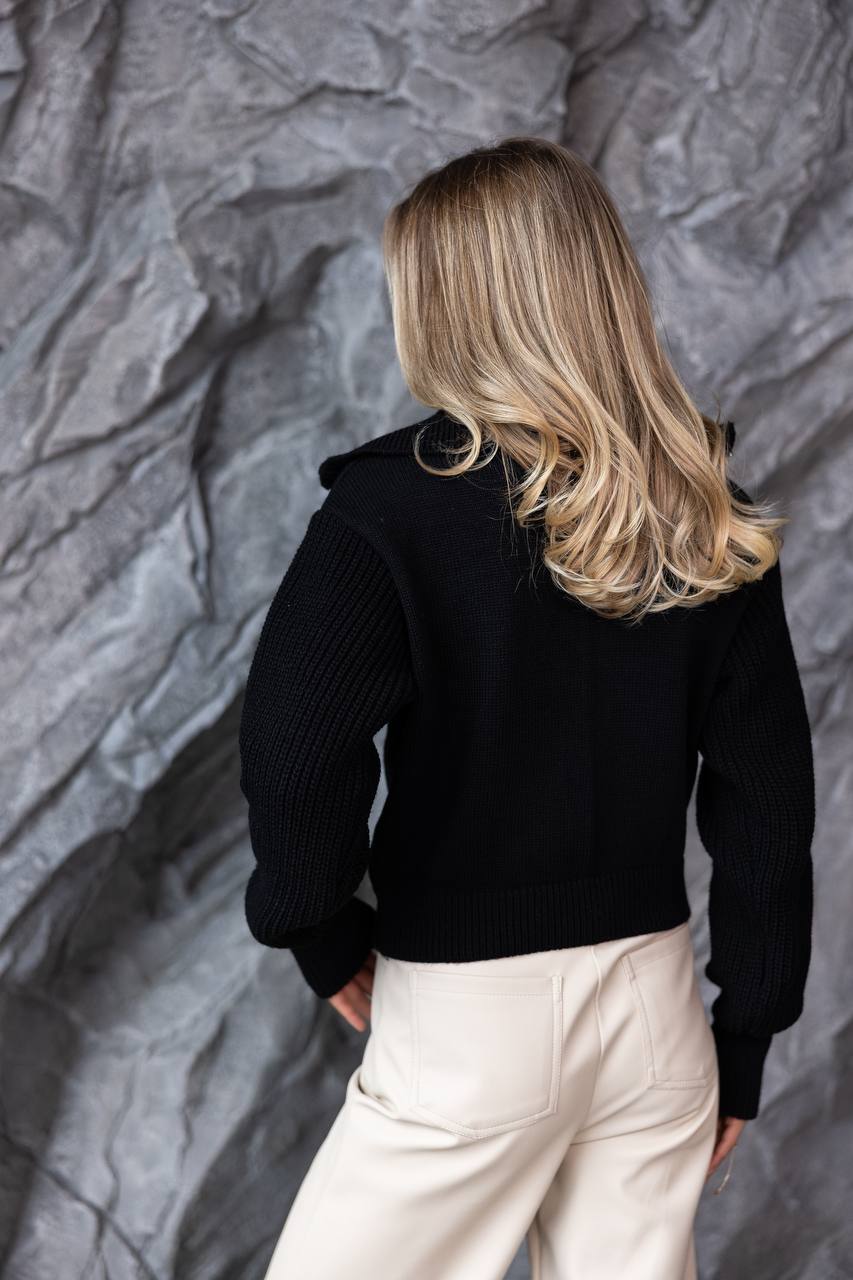 Person wearing a black jacket and white pants against a textured gray wall