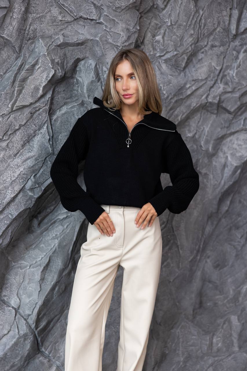 Woman wearing a black sweater and white pants against a textured gray wall