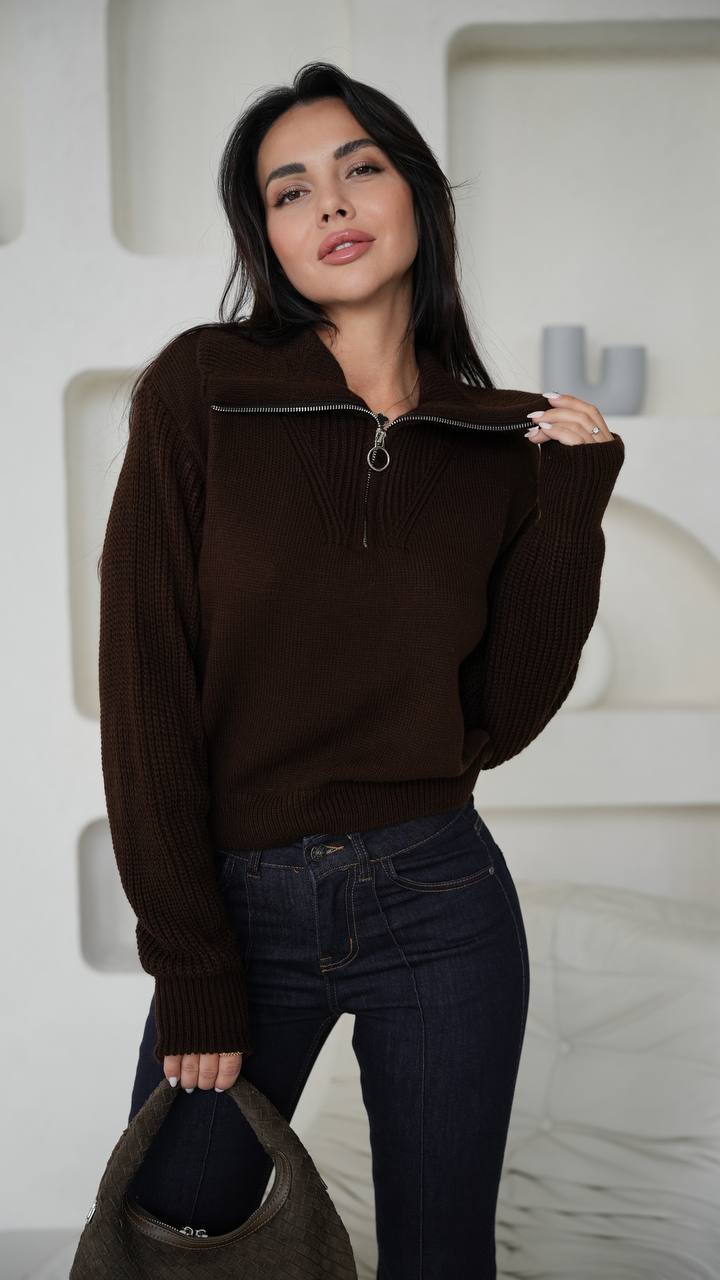 Woman wearing a brown zip-up sweater and dark jeans against a white textured wall.