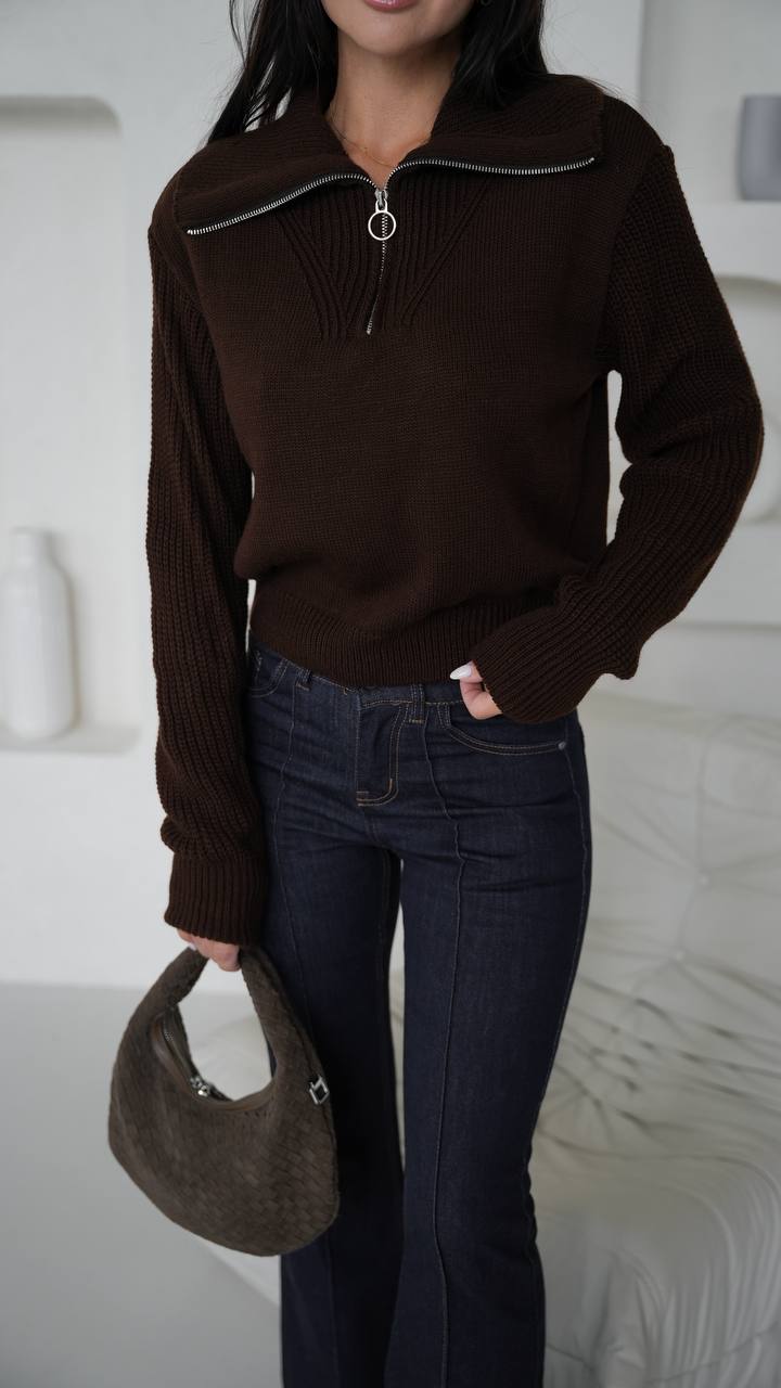 Person wearing a brown sweater and dark jeans holding a brown handbag indoors.