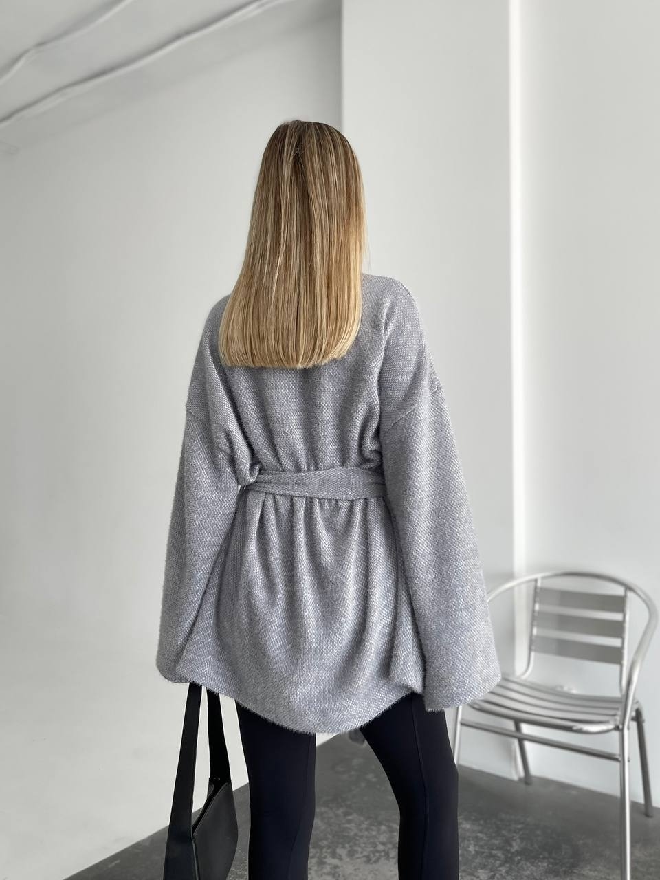 Person wearing a gray robe standing in a minimalistic room with a chair.