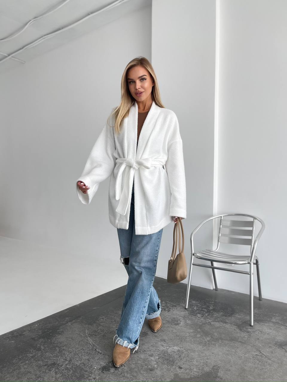 Woman wearing a white robe in a minimalistic room with a chair and bag.