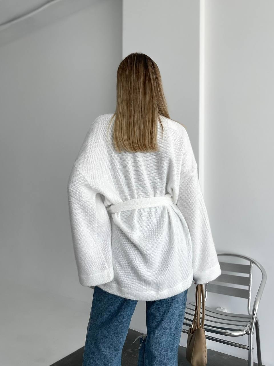 Person wearing a white robe standing in a minimalistic room with a chair and handbag.