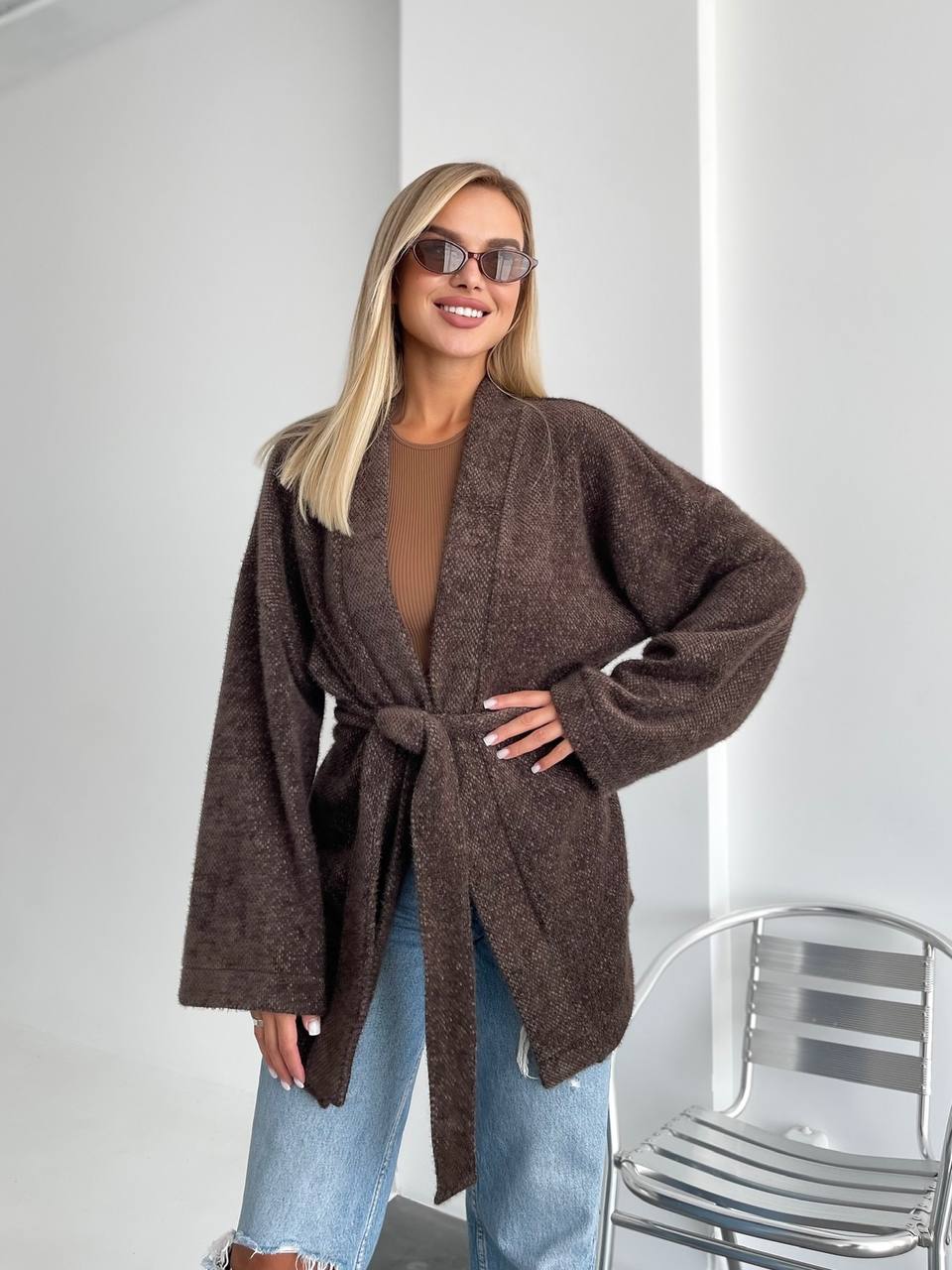 Woman wearing a brown cardigan, sunglasses, and blue jeans indoors.