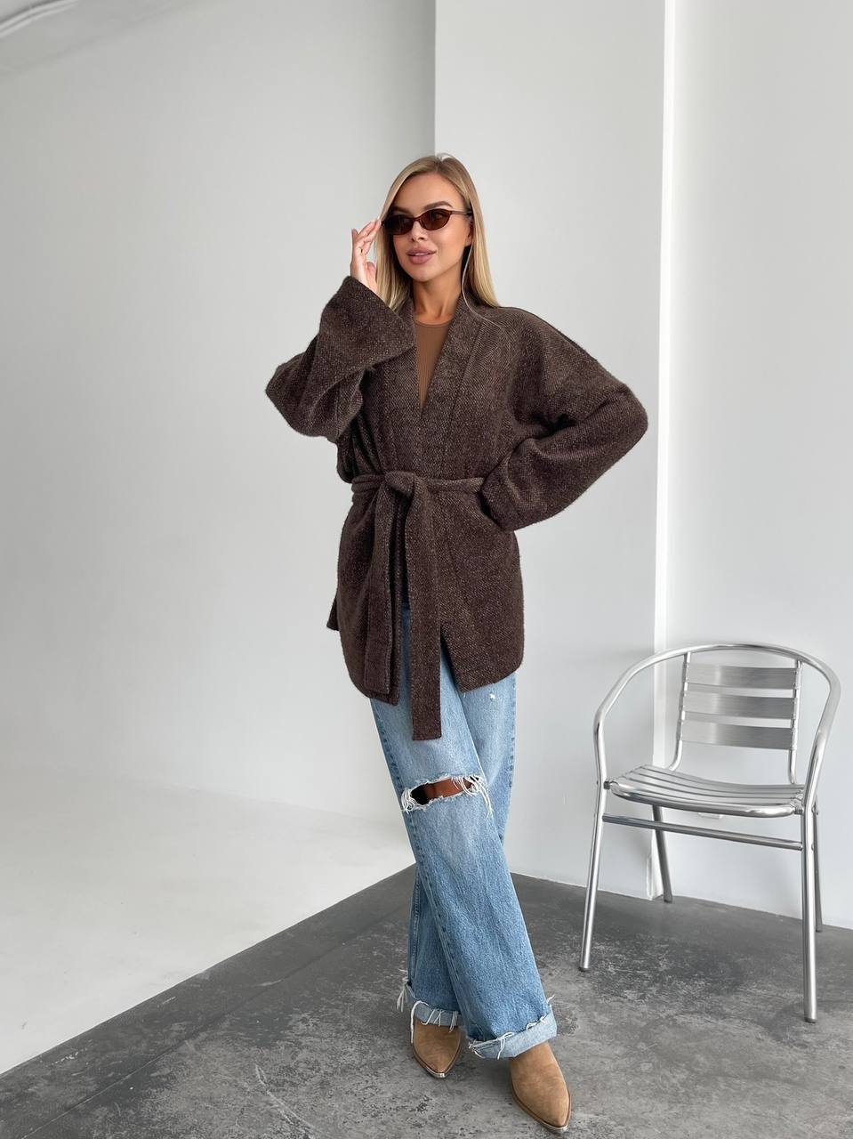 Woman wearing a brown coat and blue jeans standing in a minimalistic room with a chair.