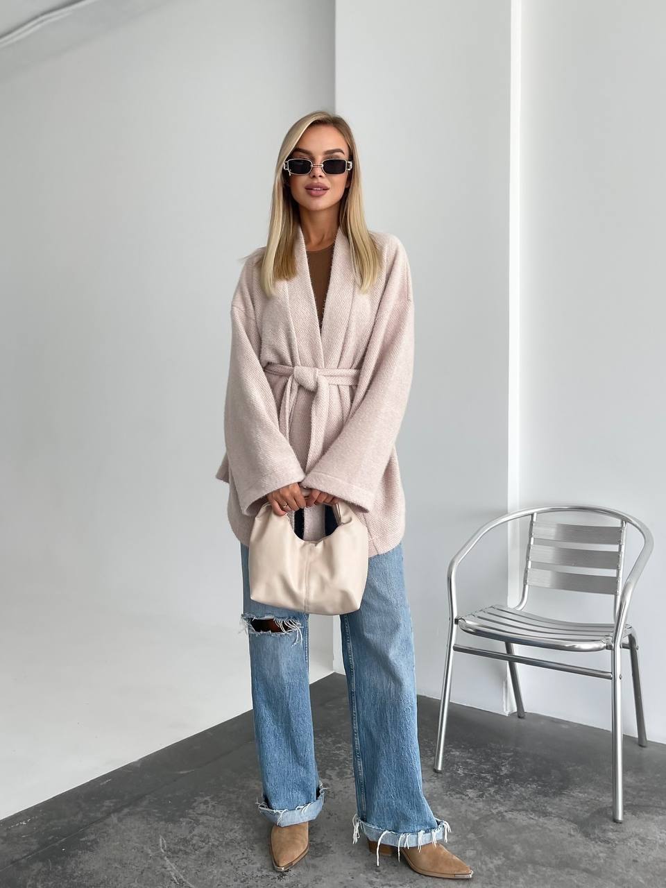 Woman wearing a light pink coat and blue jeans, holding a beige bag in a minimalistic setting.