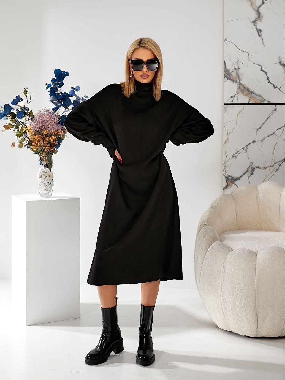 Woman wearing a black dress and boots in a modern interior setting with a vase of flowers and a chair.