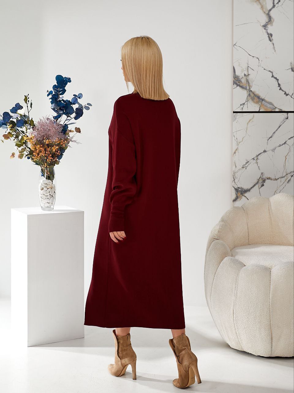 Woman in a burgundy dress standing in a modern living room with a vase of flowers and a white sofa.