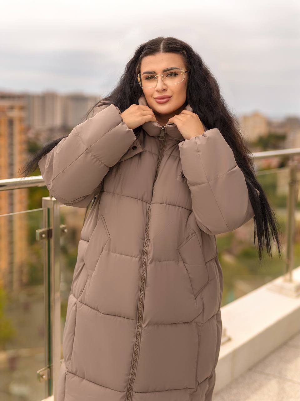 Woman wearing a beige puffer jacket on a balcony with a cityscape background