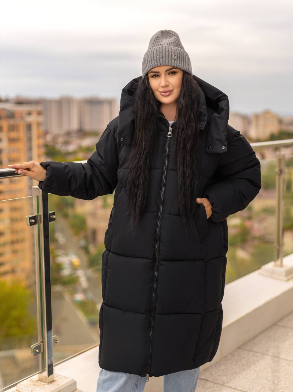 Woman wearing a black puffer coat and gray beanie standing on a balcony with a cityscape background.