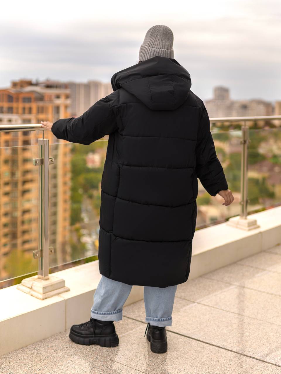 Person wearing a long black puffer coat on a rooftop with a cityscape view.