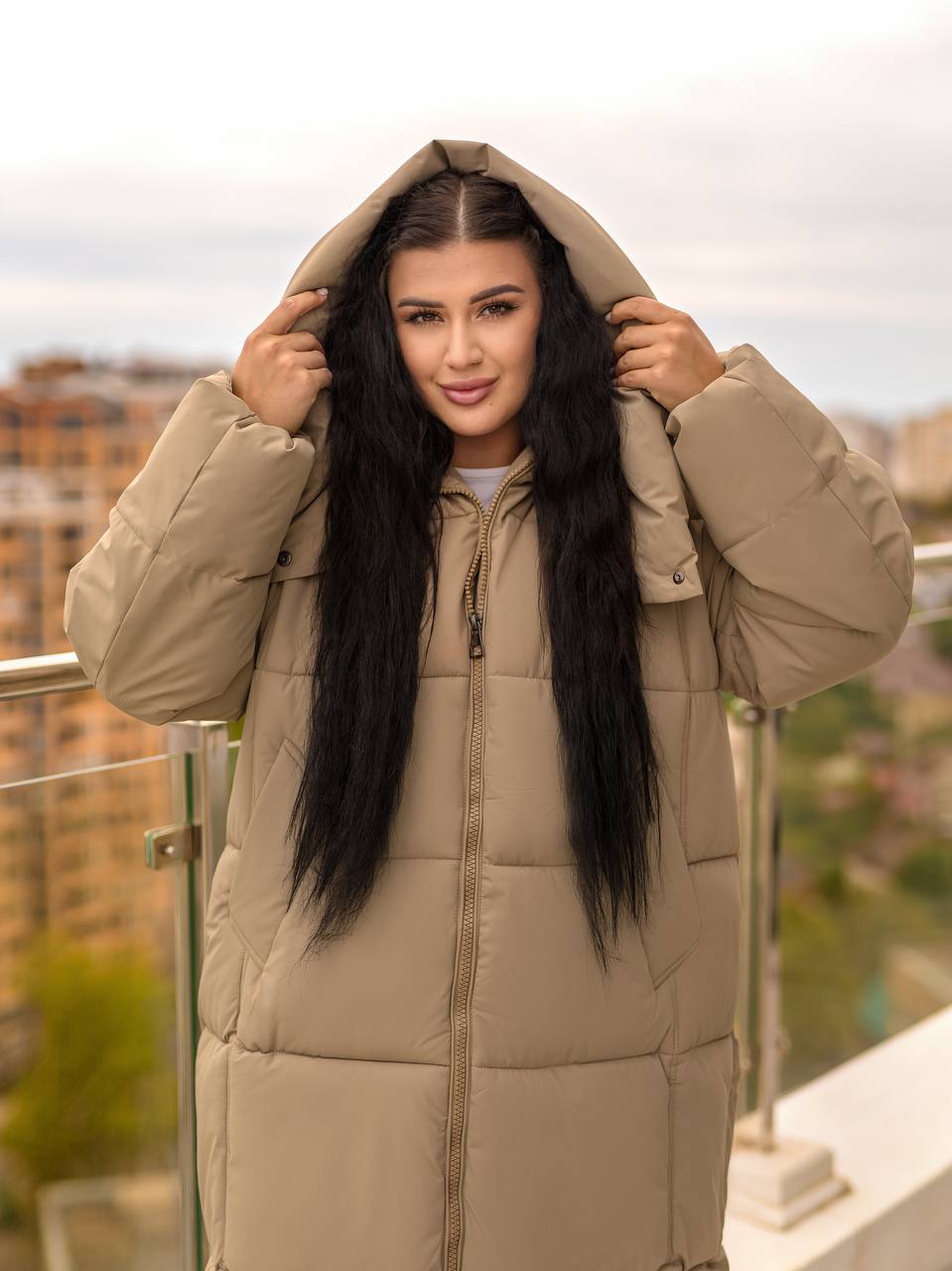 Woman wearing a beige puffer coat on a balcony with a blurred cityscape background