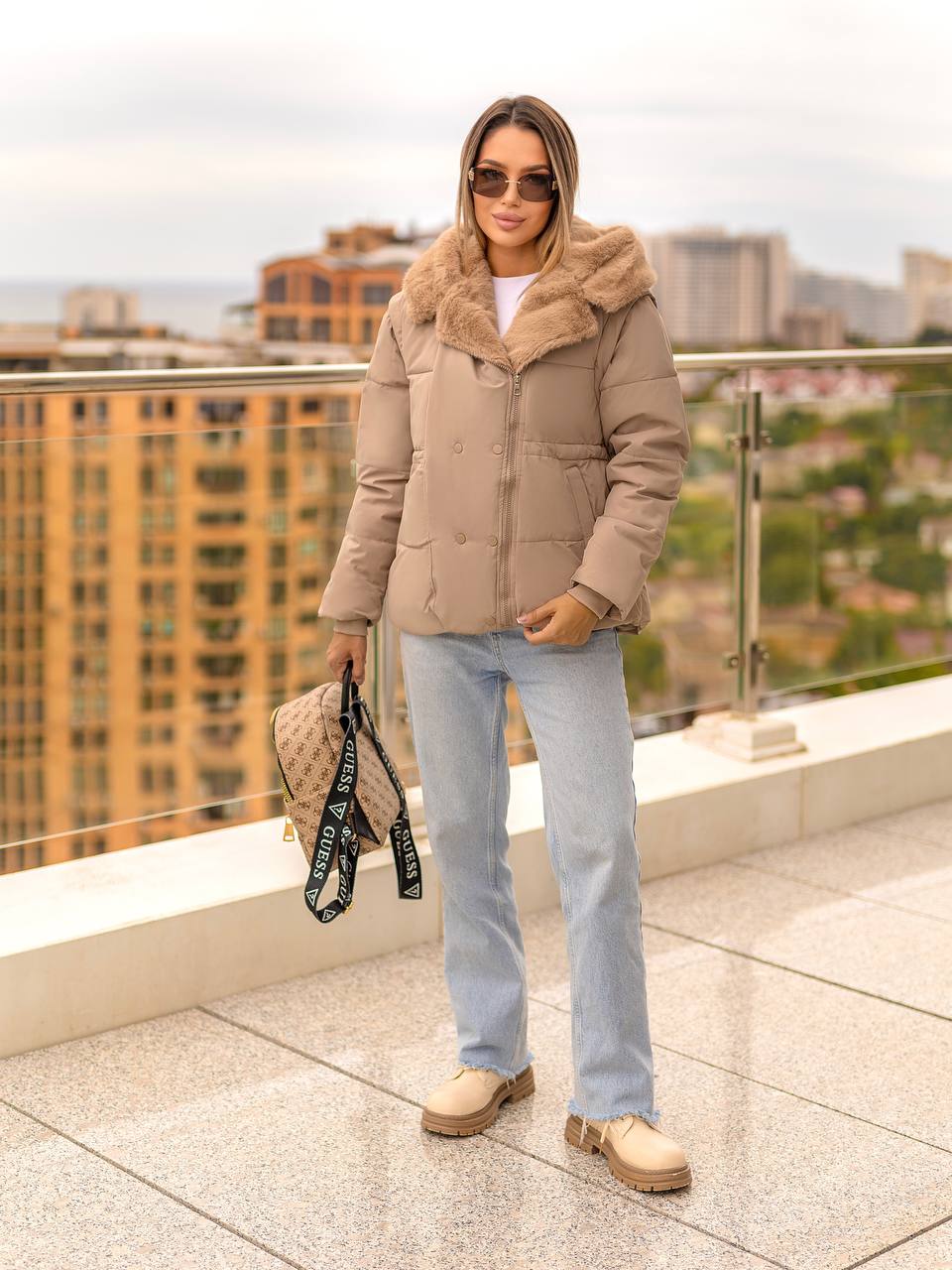 Woman wearing a beige puffer jacket with fur hood on a rooftop with cityscape background