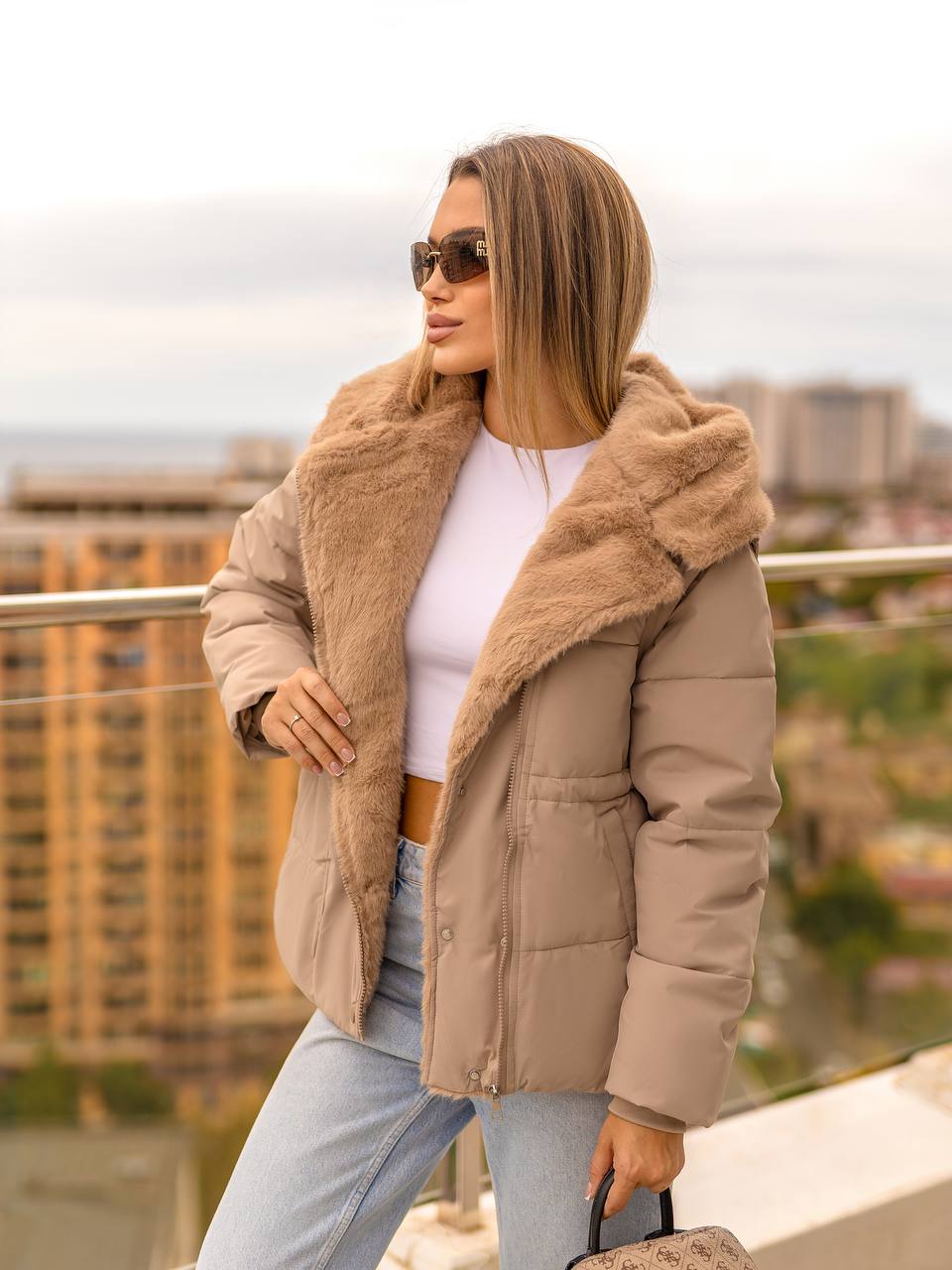 Woman wearing a beige puffer jacket with fur trim on a rooftop with cityscape background