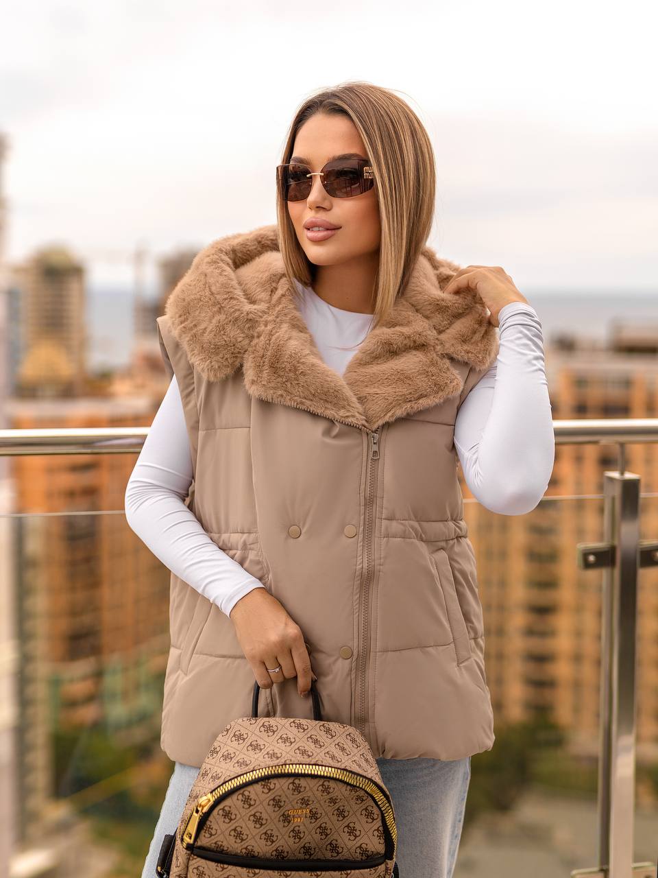Woman wearing a beige puffer vest with fur collar on a balcony with cityscape background