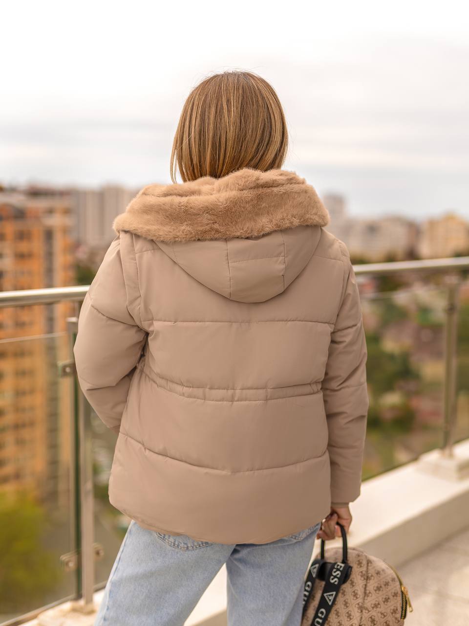 Person wearing a beige puffer jacket with fur hood on a balcony overlooking a cityscape.