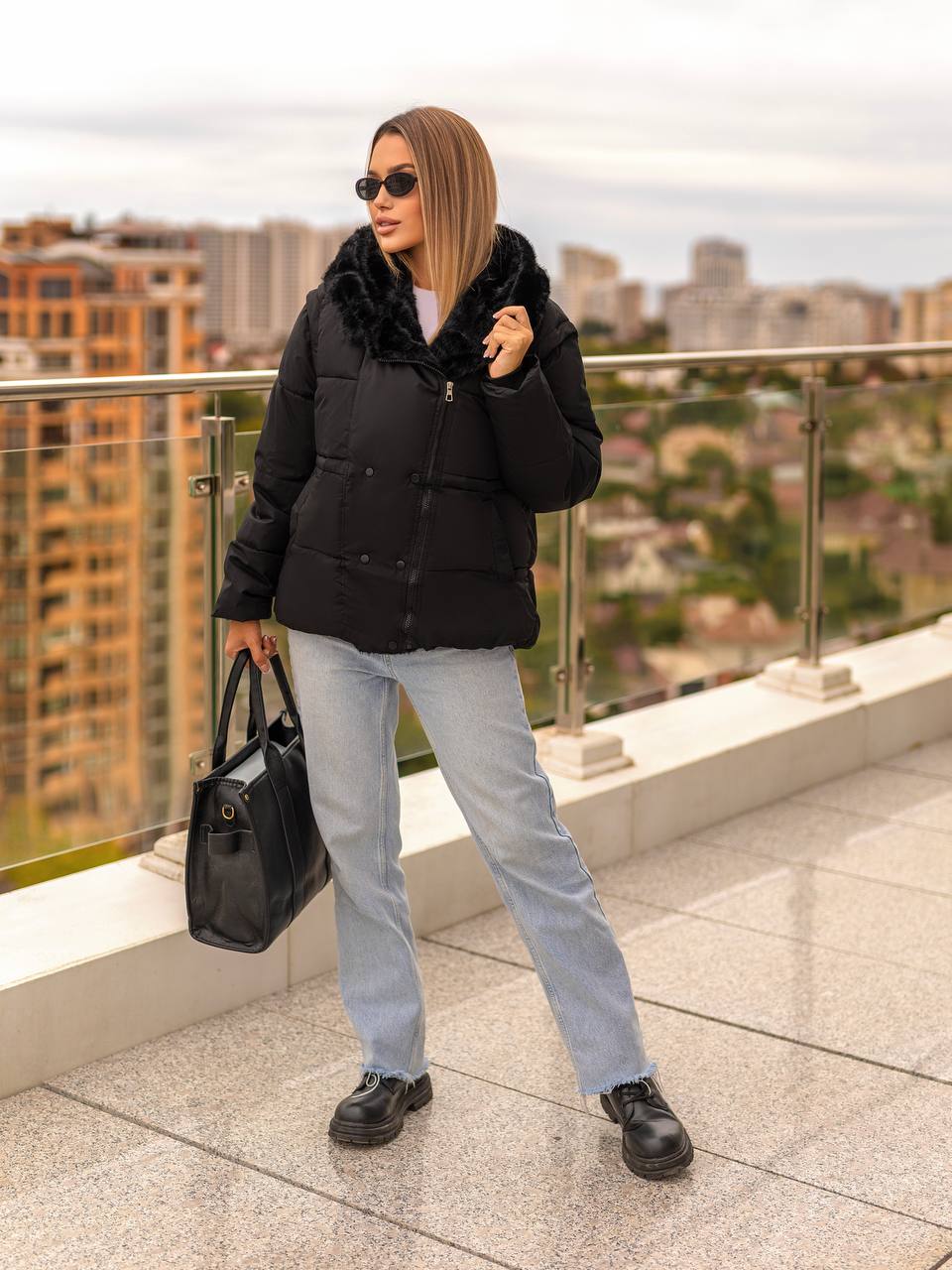 Woman in black puffer jacket with fur hood, light blue jeans, and black shoes standing on a rooftop with cityscape background.