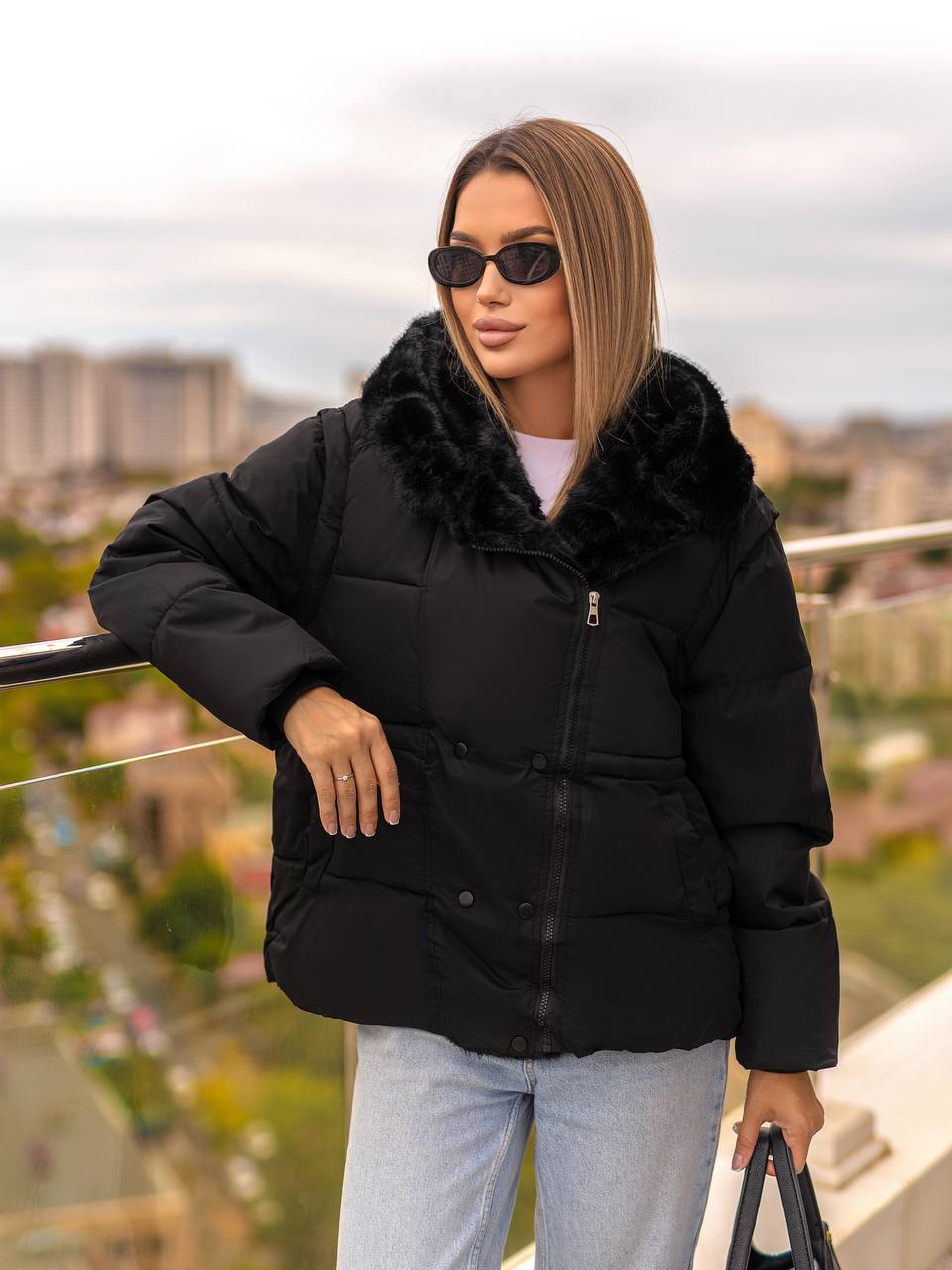 Woman wearing a black puffer jacket with fur hood on a balcony with a cityscape background