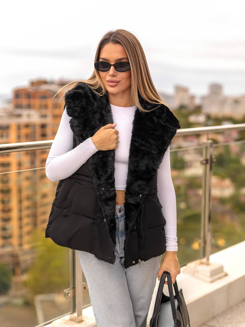 Woman wearing a black fur vest on a balcony with a cityscape background
