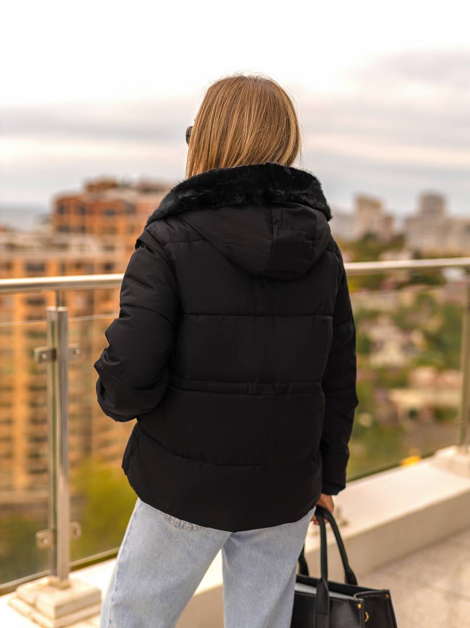 Person wearing a black puffer jacket with fur hood, standing on a balcony overlooking a cityscape.