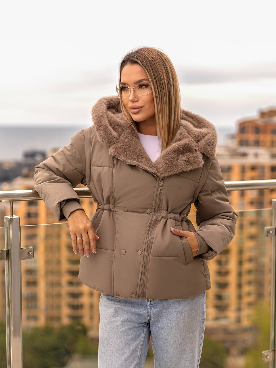 Woman wearing a beige puffer jacket with fur trim on a city balcony.