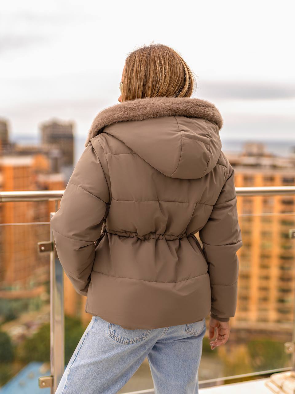 Person wearing a brown puffer jacket with a fur-lined hood on a rooftop with cityscape view.