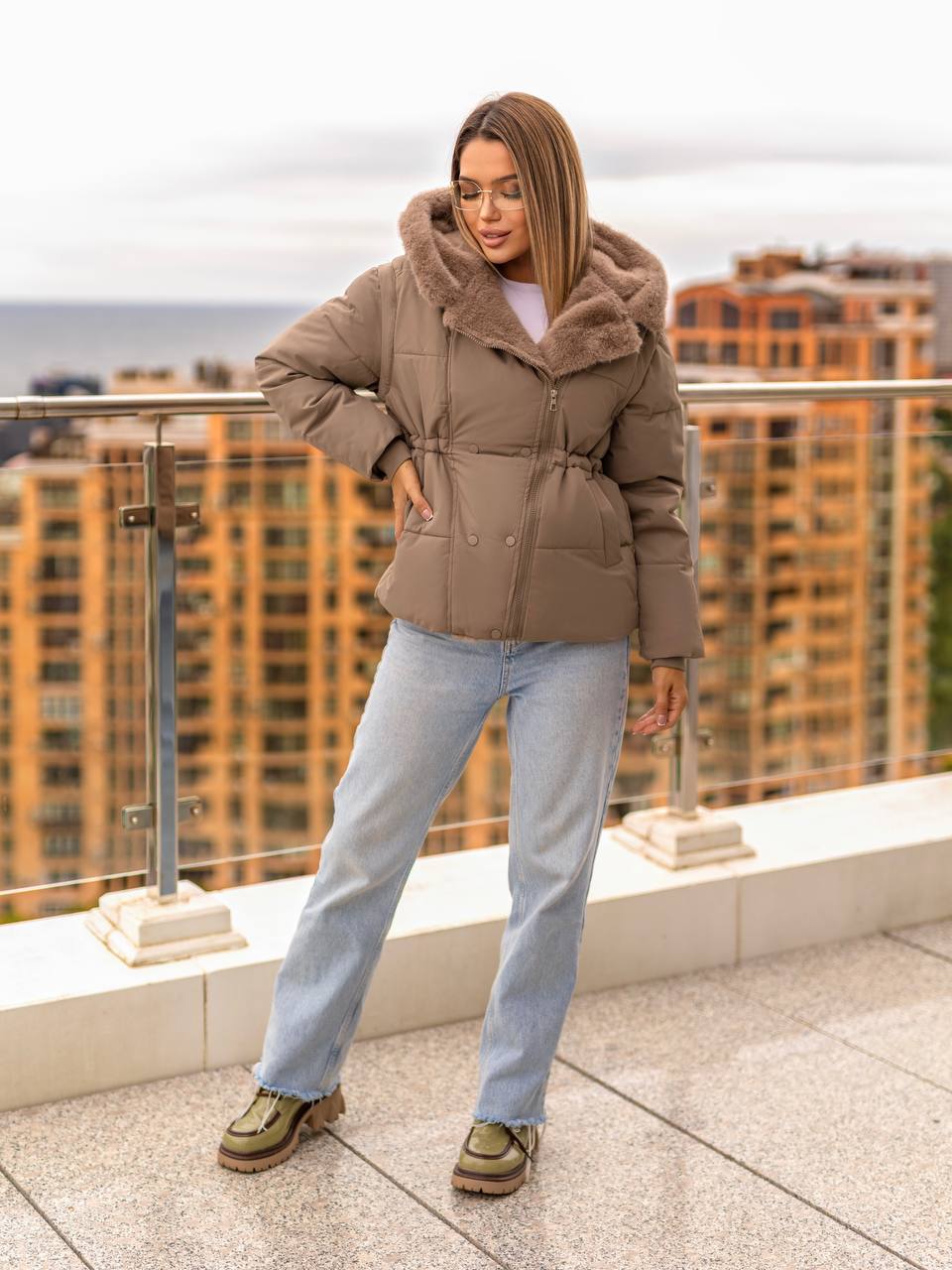 Woman wearing a brown puffer jacket on a rooftop with cityscape background