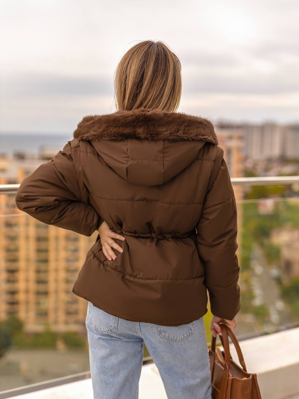 Person wearing a brown puffer jacket with fur collar on a balcony with cityscape view