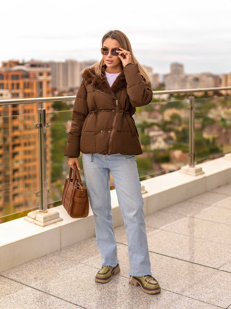 Woman in a brown puffer jacket and light blue jeans standing on a rooftop with a cityscape background.
