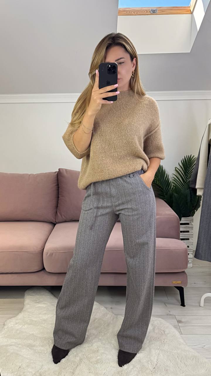 Person taking a mirror selfie wearing a beige sweater and gray pants in a living room.