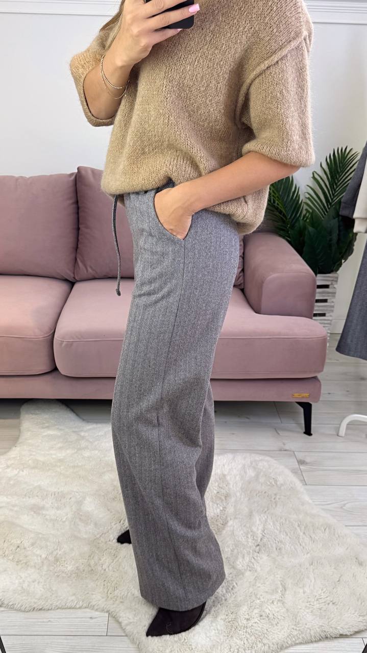 Person wearing a beige sweater and gray pants standing in a living room.
