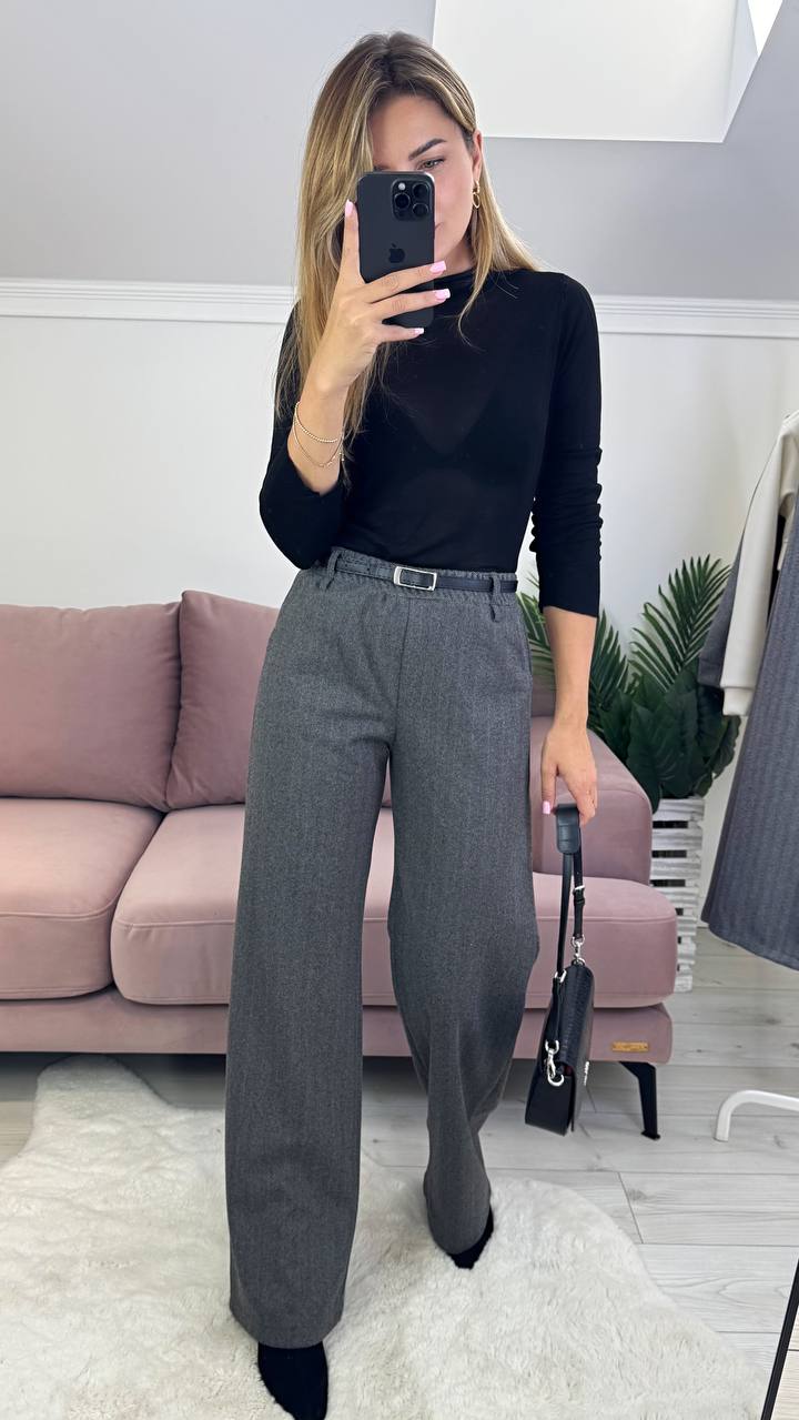 Woman taking a mirror selfie wearing a black top and gray pants in a room with a pink couch and white walls.