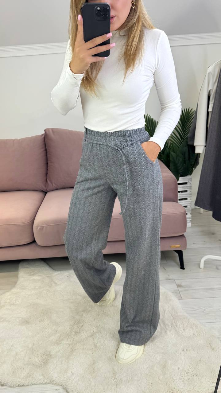 Person wearing a white top and gray pants taking a mirror selfie in a room with a couch and plant.