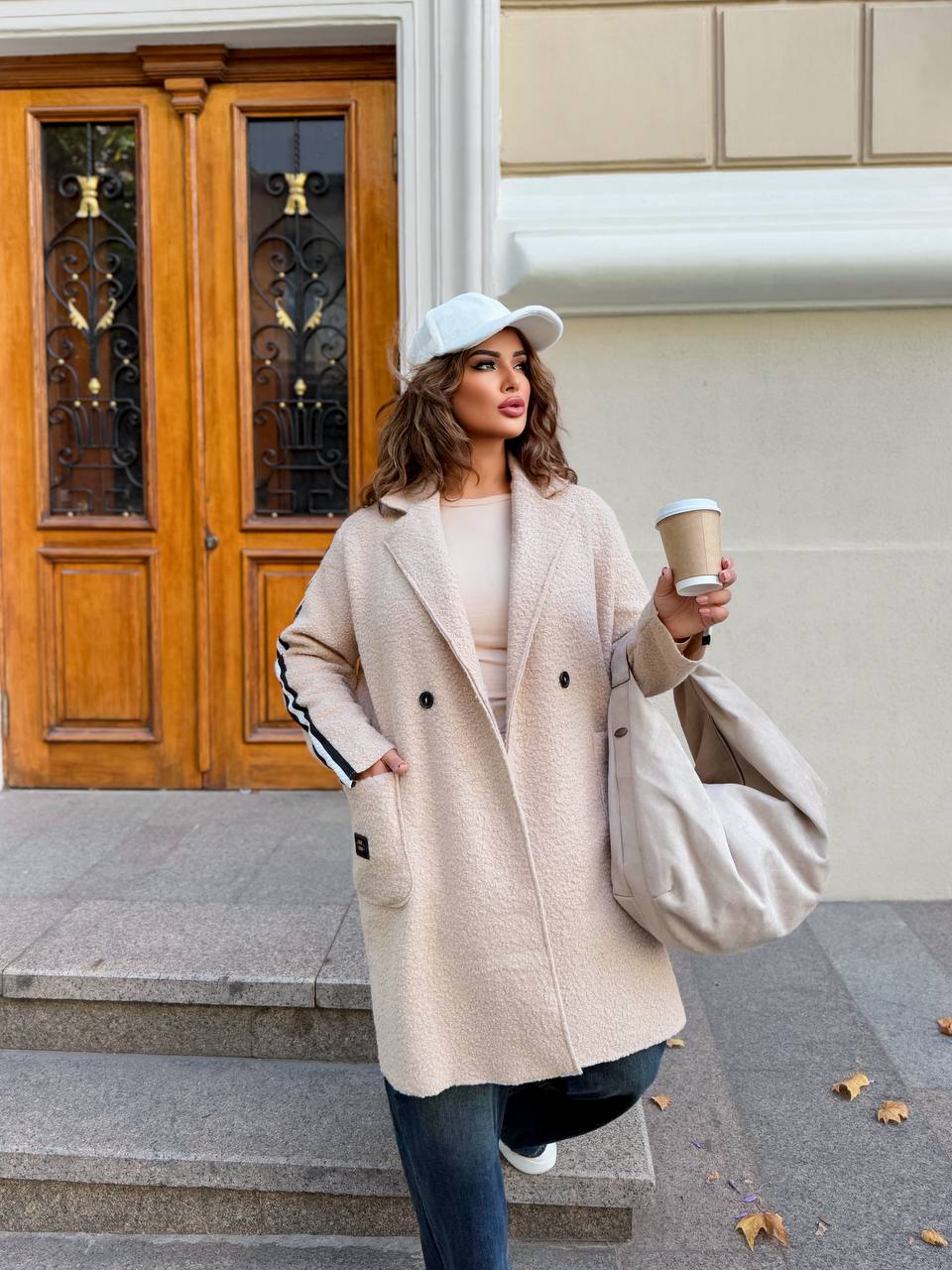 Woman in a beige coat holding a coffee cup and a bag, standing in front of a building entrance.