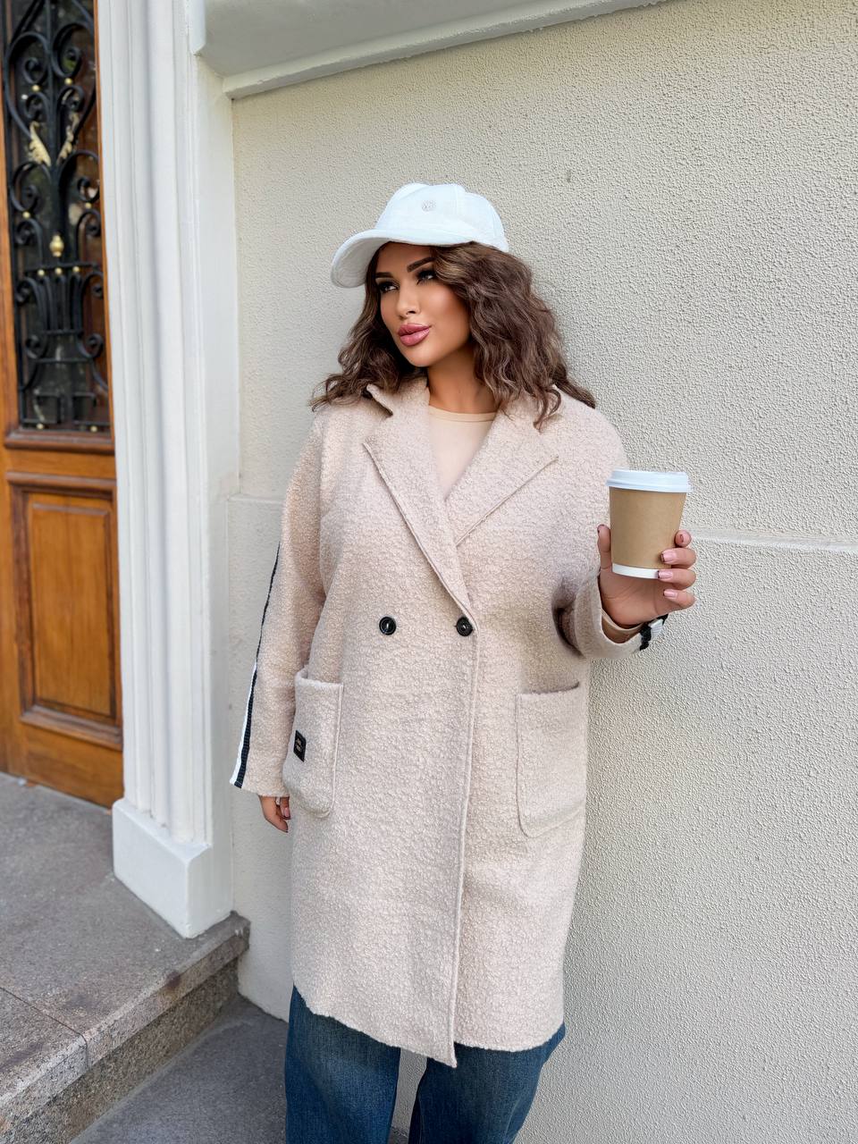 Woman in a beige coat and white cap holding a coffee cup against a light-colored wall.